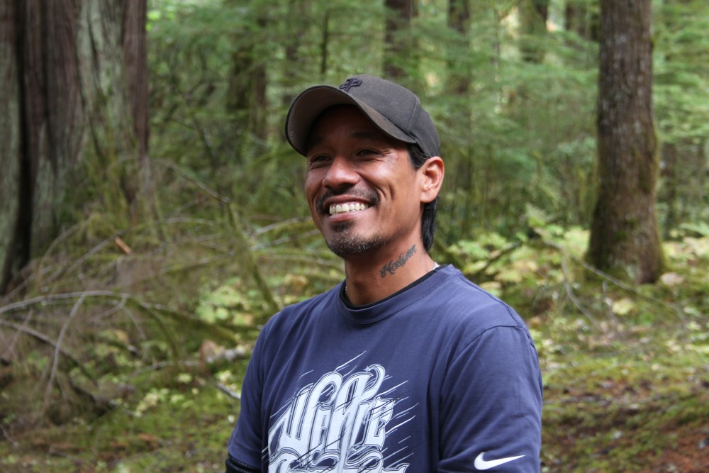 A smiling man in a blue shirt wearing a baseball cap stands in a grove of trees.