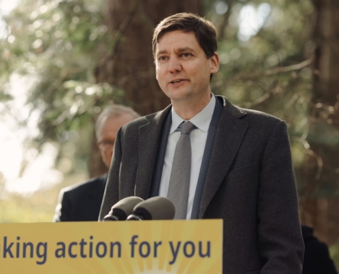 Premier David Eby stands at a yellow podium that reads, "Taking action for you," with trees in the background.